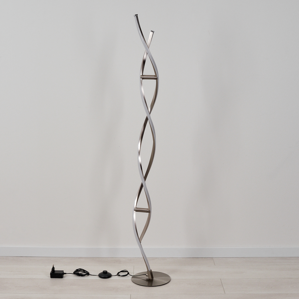 Eine moderne silberne Stehlampe mit gewundenem Design. Sie steht auf einem runden Sockel, mit einem schwarzen Netzkabel und einem Schalter.