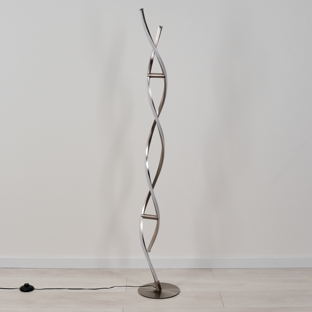 Eine moderne, silberne Stehlampe. Sie hat ein wirbelndes Design, einen kreisförmigen Sockel und ein schwarzes Netzkabel. Sie steht auf einem hellen Holzboden.