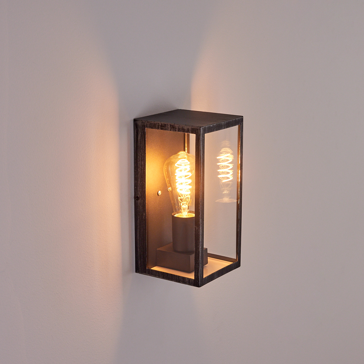 Eine wandmontierte, rechteckige Außenleuchte mit einer leuchtenden Edison-Glühbirne in einem Glas- und Metallrahmen. Die Wand ist hellgrau.