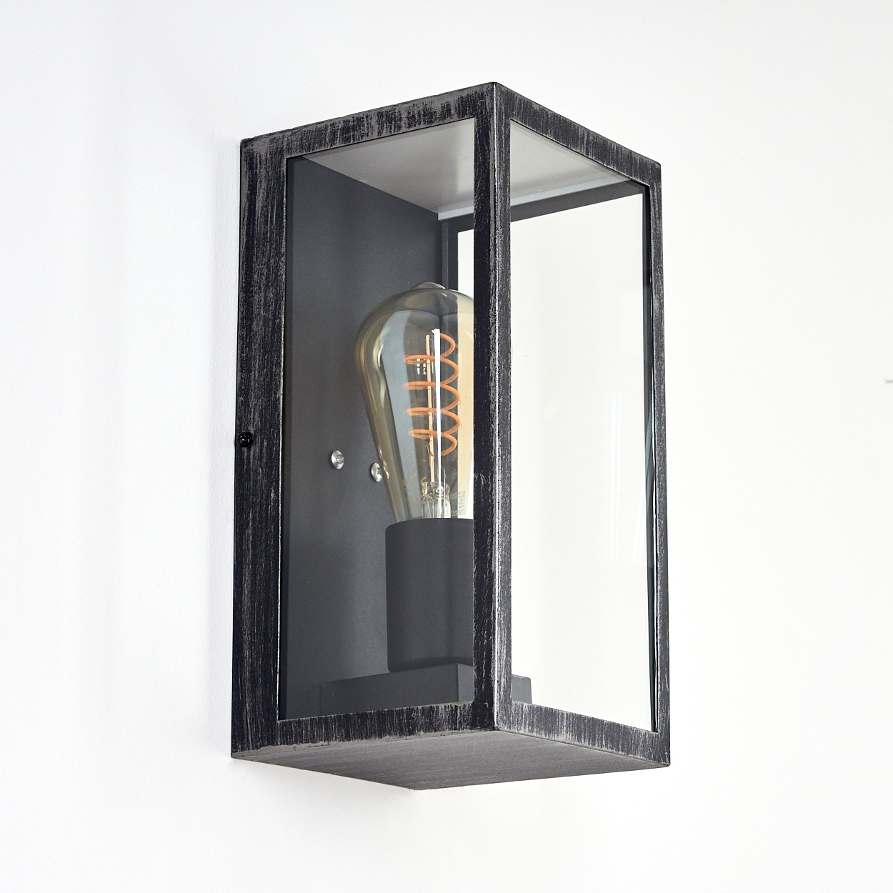 Eine rechteckige Wandleuchte mit einer Vintage-Glühbirne, umhüllt von einem dunklen Metallrahmen mit Glasscheiben, an einer weißen Wand montiert.