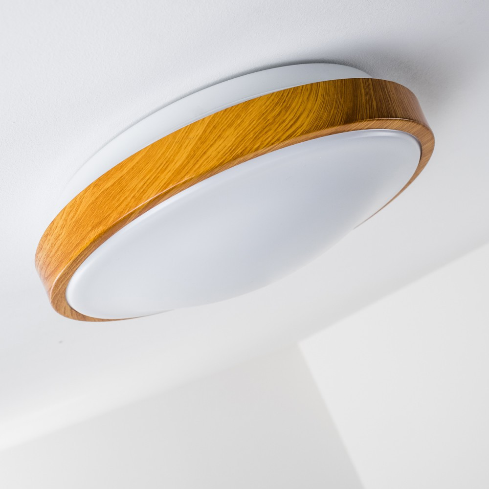 Eine moderne Deckenleuchte mit weißer, runder Lichtquelle und einem gebogenen Holzrahmen. Der Hintergrund ist eine weiße Decke.