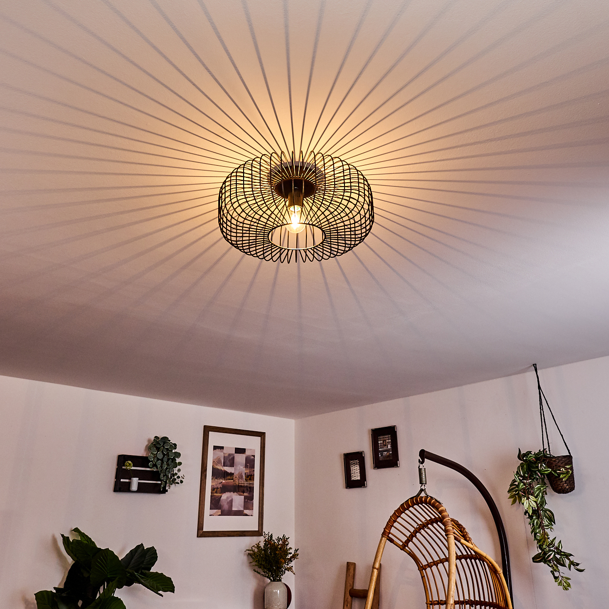 Eine schwarze, kreisförmige Drahtgestell-Leuchte wirft radiale Schatten auf die weiße Decke. Darunter ist ein Raum mit Dekoration sichtbar.