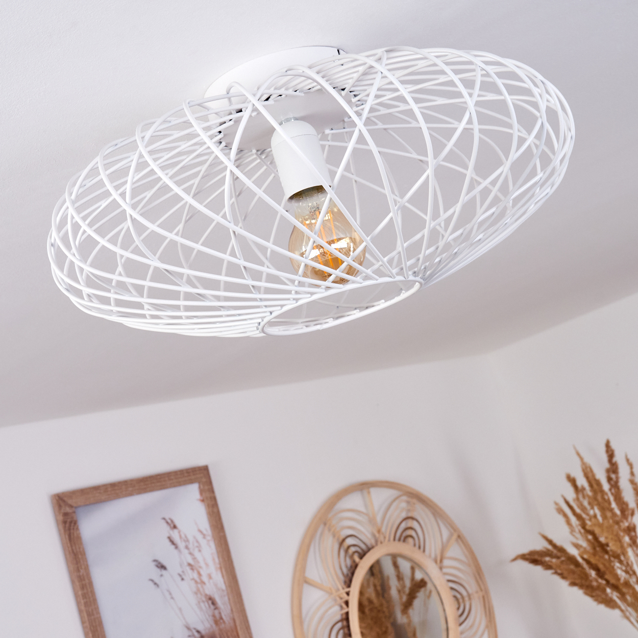 Eine weiße, ovale Deckenlampe mit sichtbarer Glühbirne. Darunter sind ein Bilderrahmen und ein Rattanspiegel zu sehen.