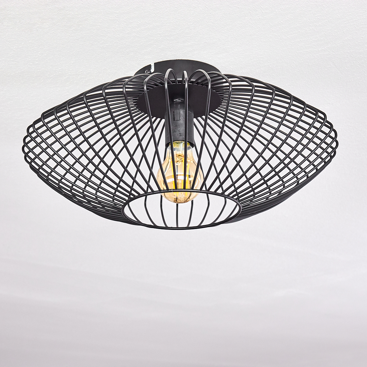 Schwarze, käfigartige Deckenlampe mit Glühbirne. Vor weißer Decke. Die Lampe hat ein geometrisches Design und die Glühbirne leuchtet.
