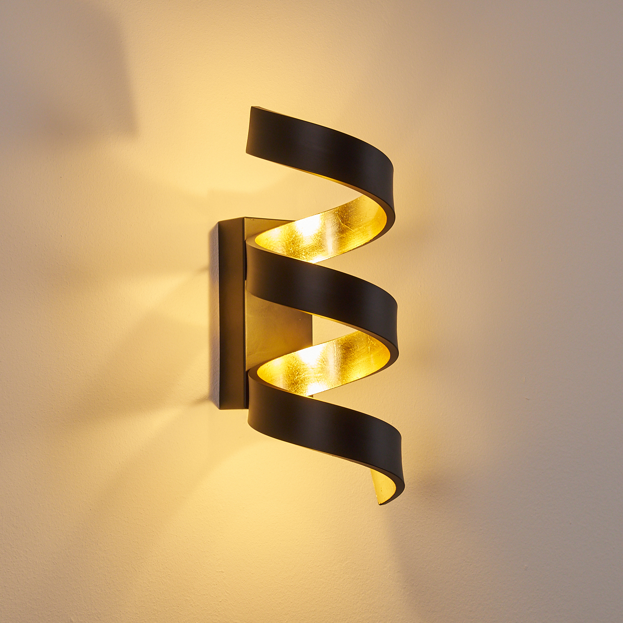 Eine Wandlampe mit Spiraldesign. Die Lampe ist schwarz mit goldener Innenseite. Die Wand ist hellbeige.
