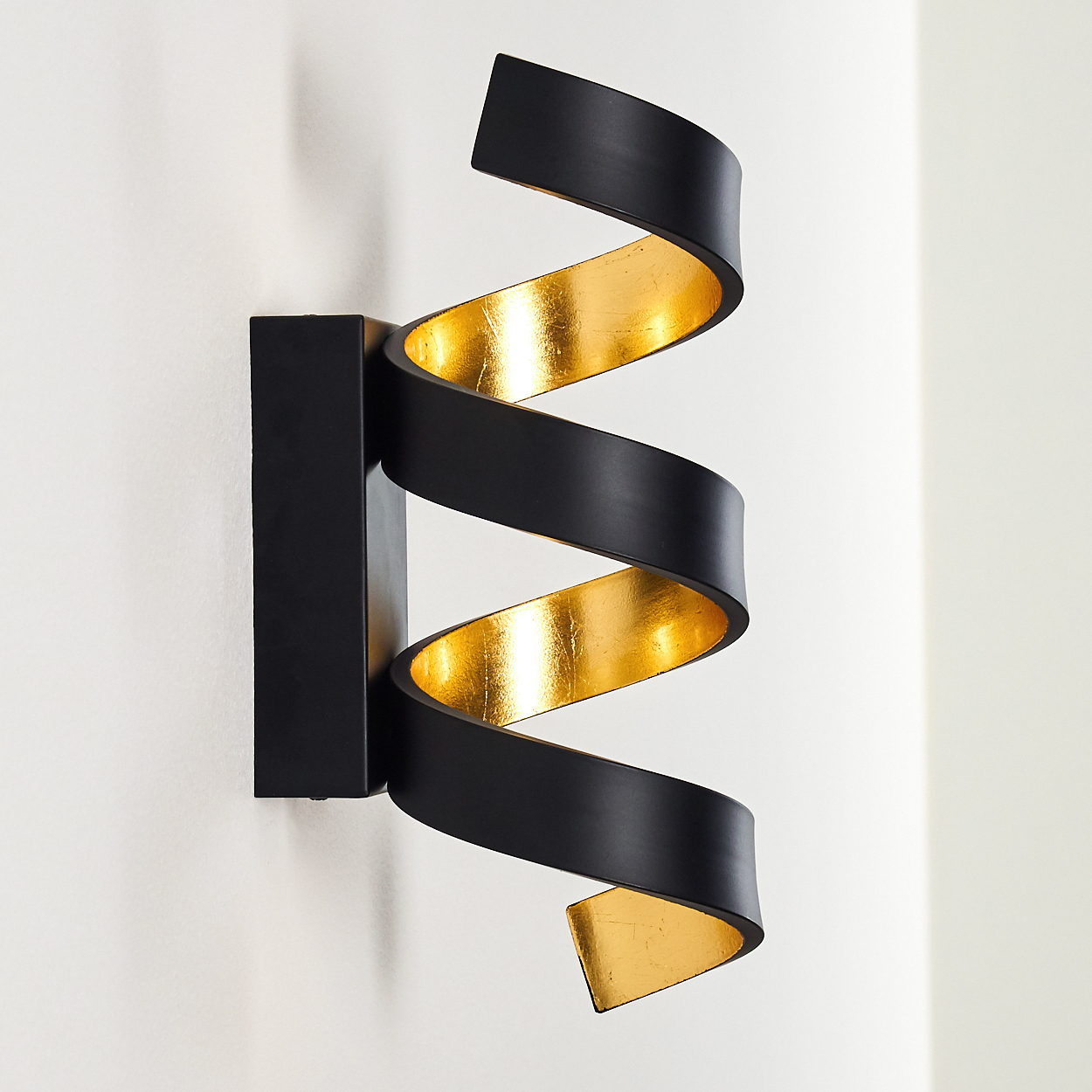 Eine Wandleuchte mit schwarzem, spiralförmigem Design und goldfarbenem Innenraum. Der Hintergrund ist eine weiße Wand.