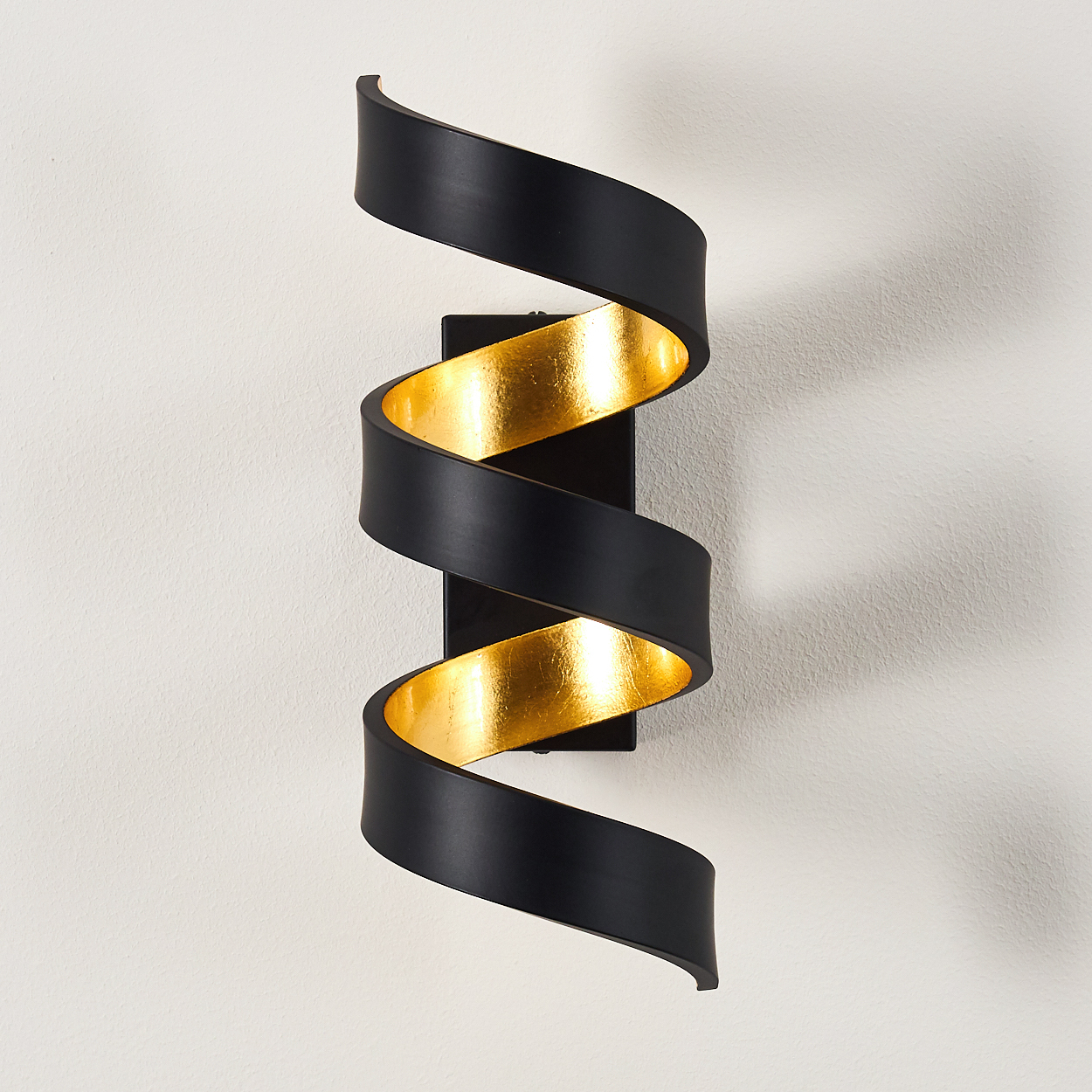 Eine schwarz-goldene Wandleuchte mit Spiraldesign an einer weißen Wand. Das Licht reflektiert von der goldenen Innenfläche.