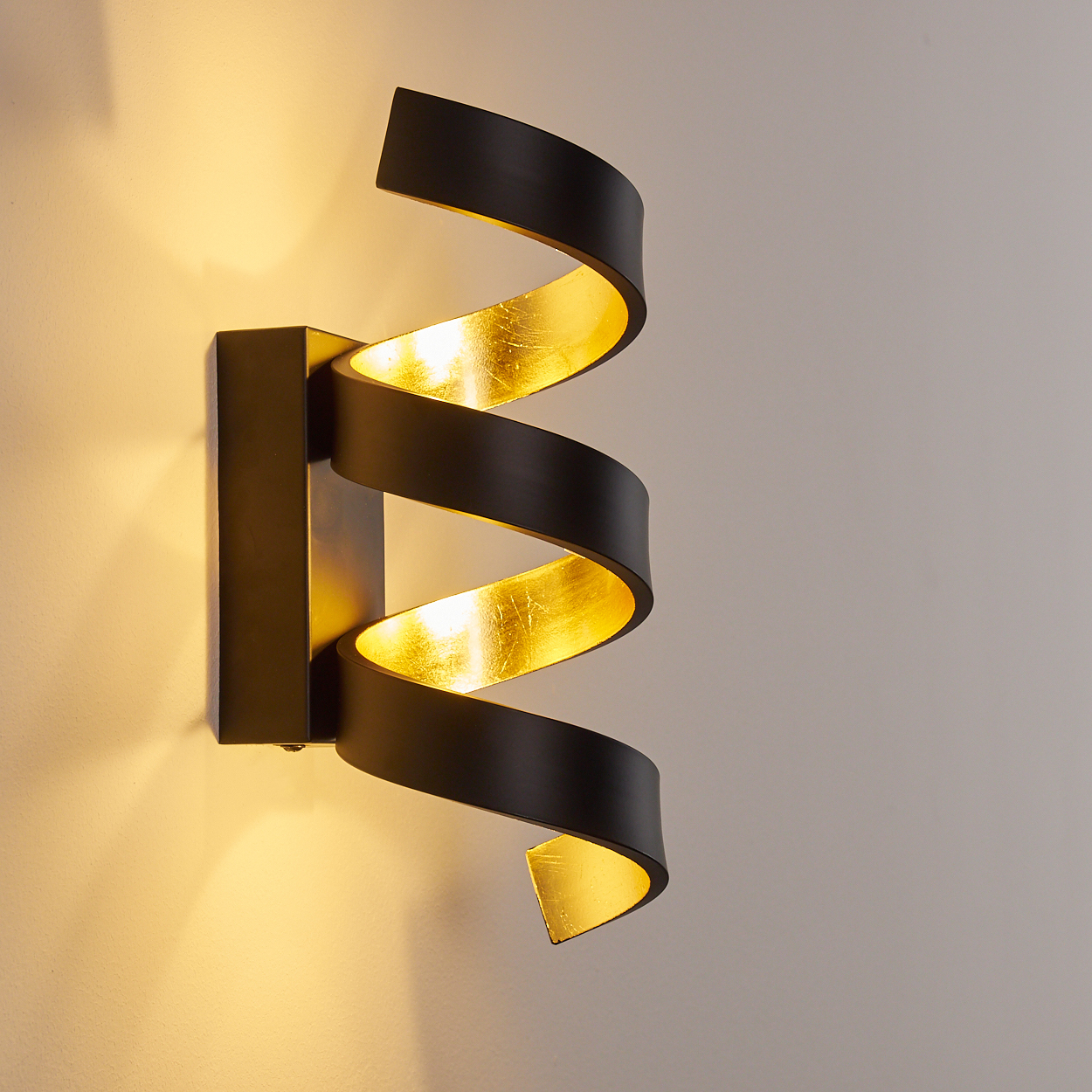 Eine schwarze Wandleuchte mit spiralförmigem Design, goldfarbenem Innenraum. Sie ist an einer hellgrauen Wand montiert.