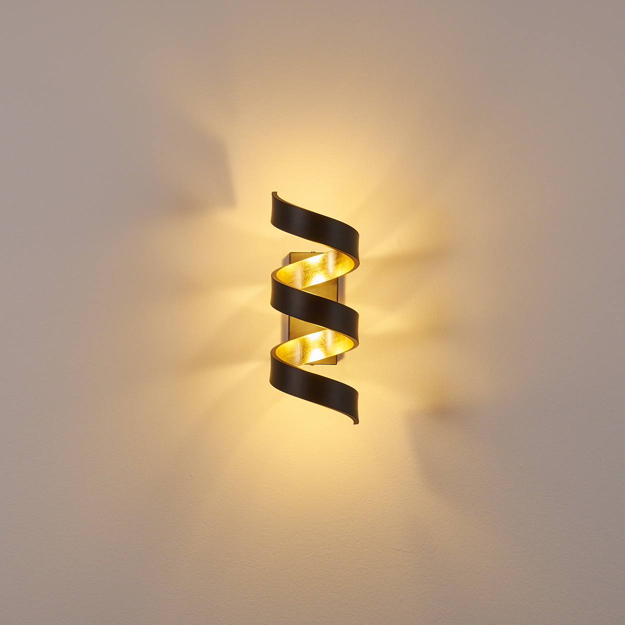 Eine Wandlampe mit Spiraldesign, schwarz und goldfarben, an einer beigefarbenen Wand. Die Lampe strahlt ein warmes, goldenes Licht aus.