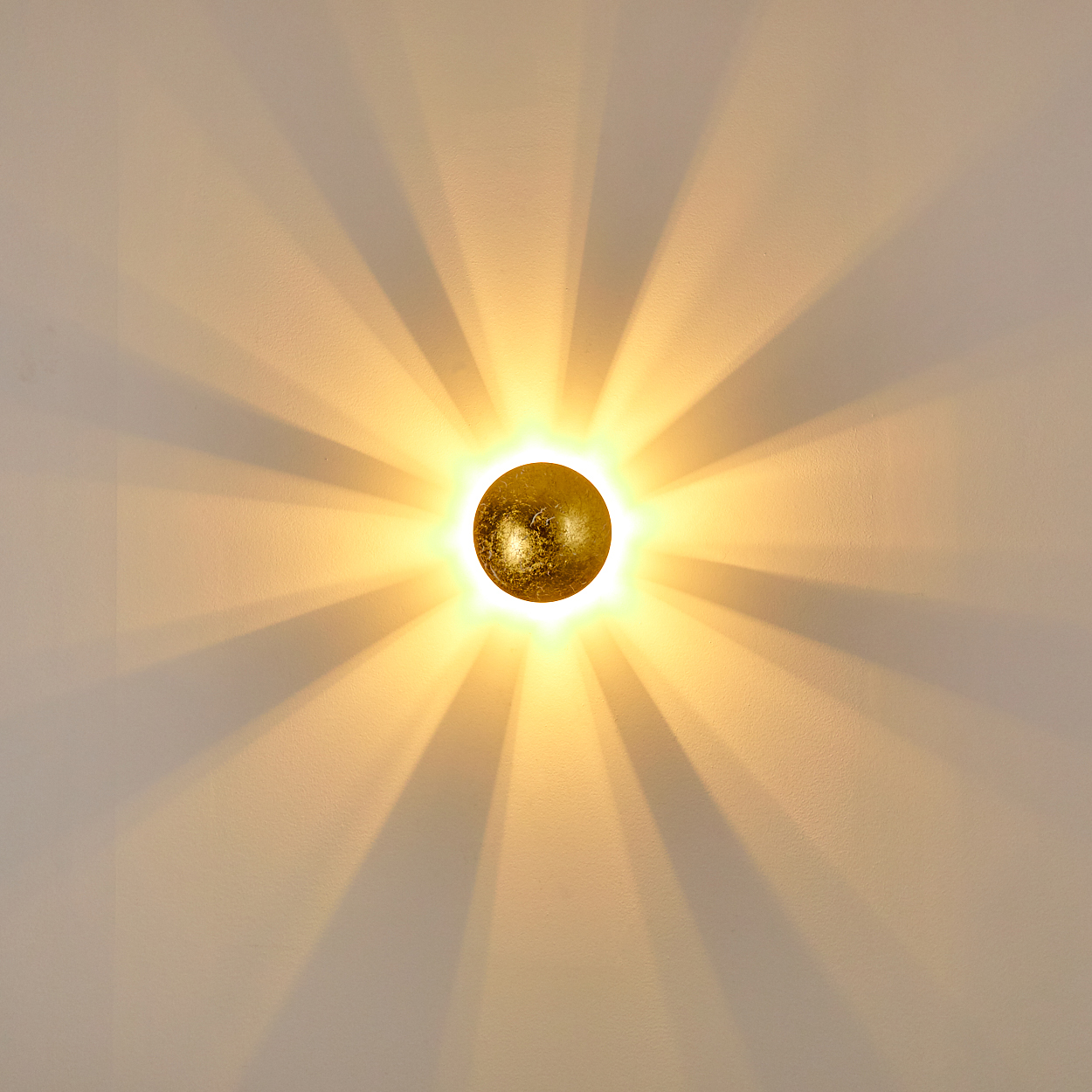 Eine runde, goldene Leuchte an einer weißen Wand. Licht strahlt aus und wirft Schatten.