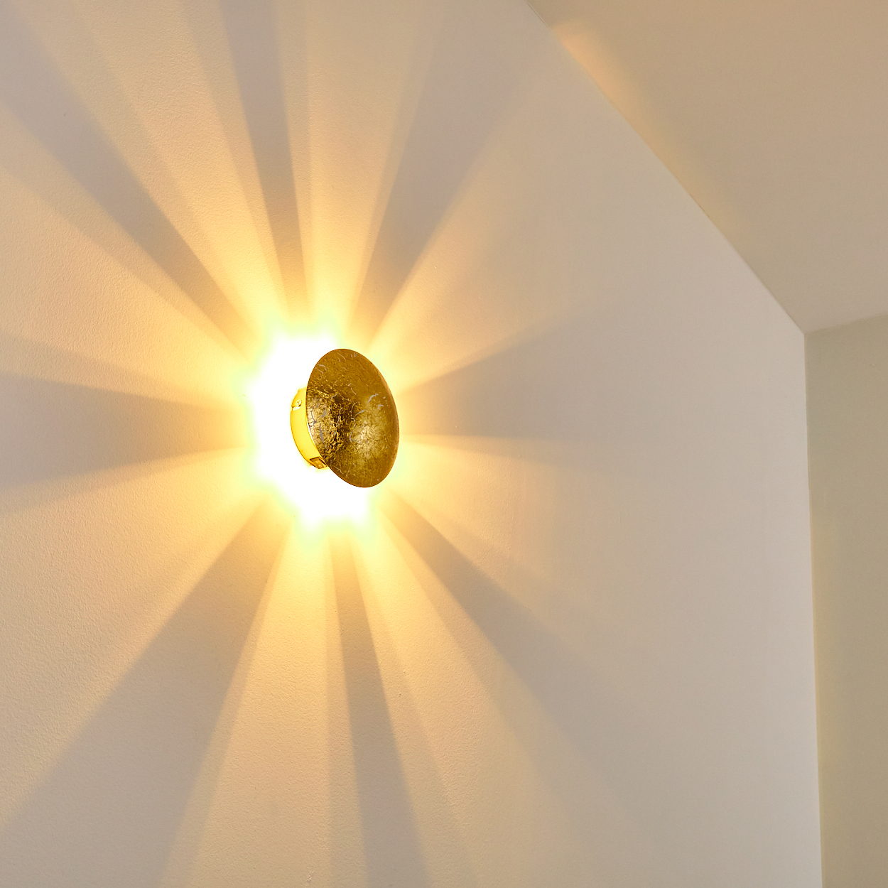 Eine goldene Wandlampe strahlt Licht mit radialen Schatten auf eine weiße Wand. Die Lampe hat eine strukturierte Oberfläche.