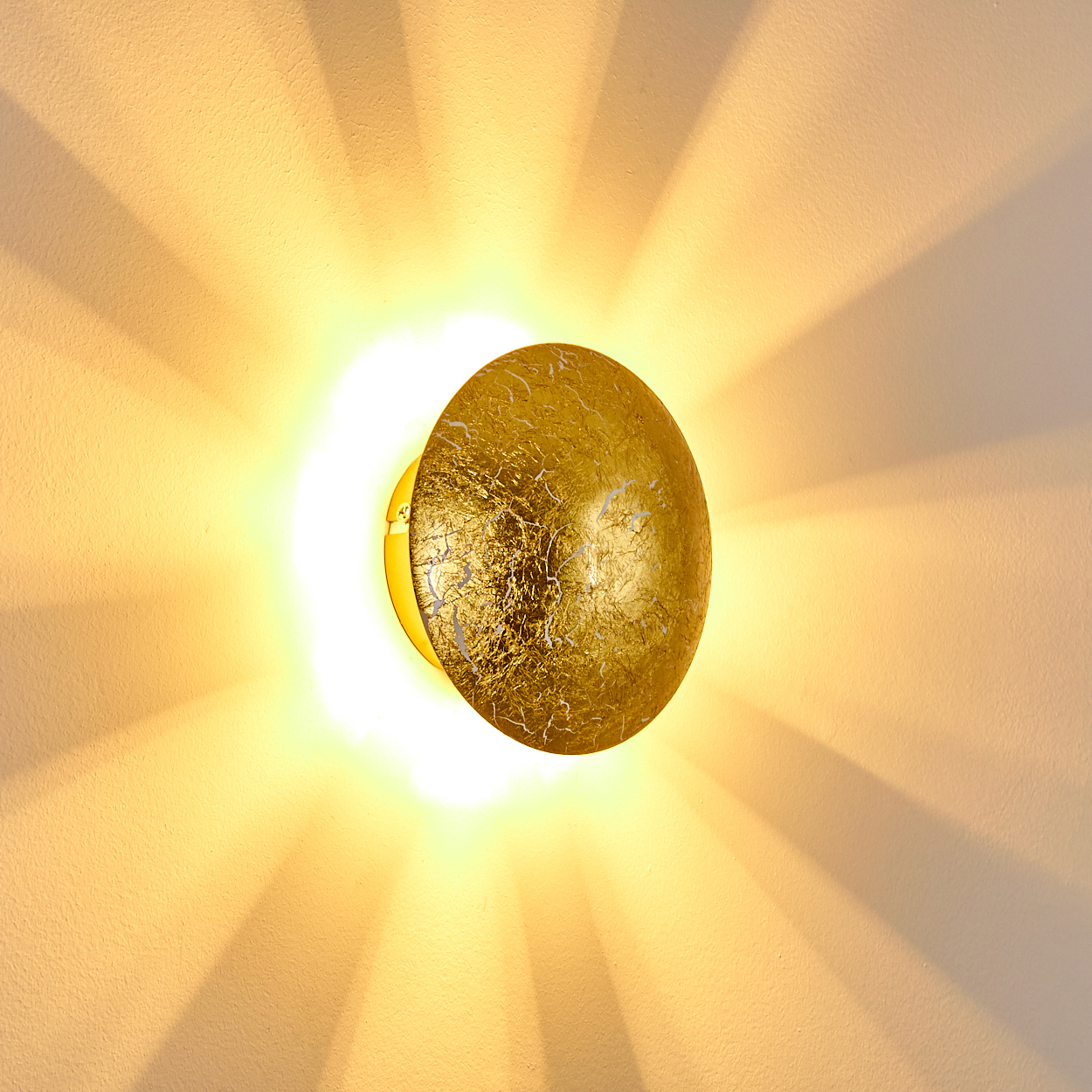 Eine goldene, strukturierte Wandleuchte, die helles Licht ausstrahlt. Lichtstrahlen strahlen auf der beigefarbenen Wand aus.