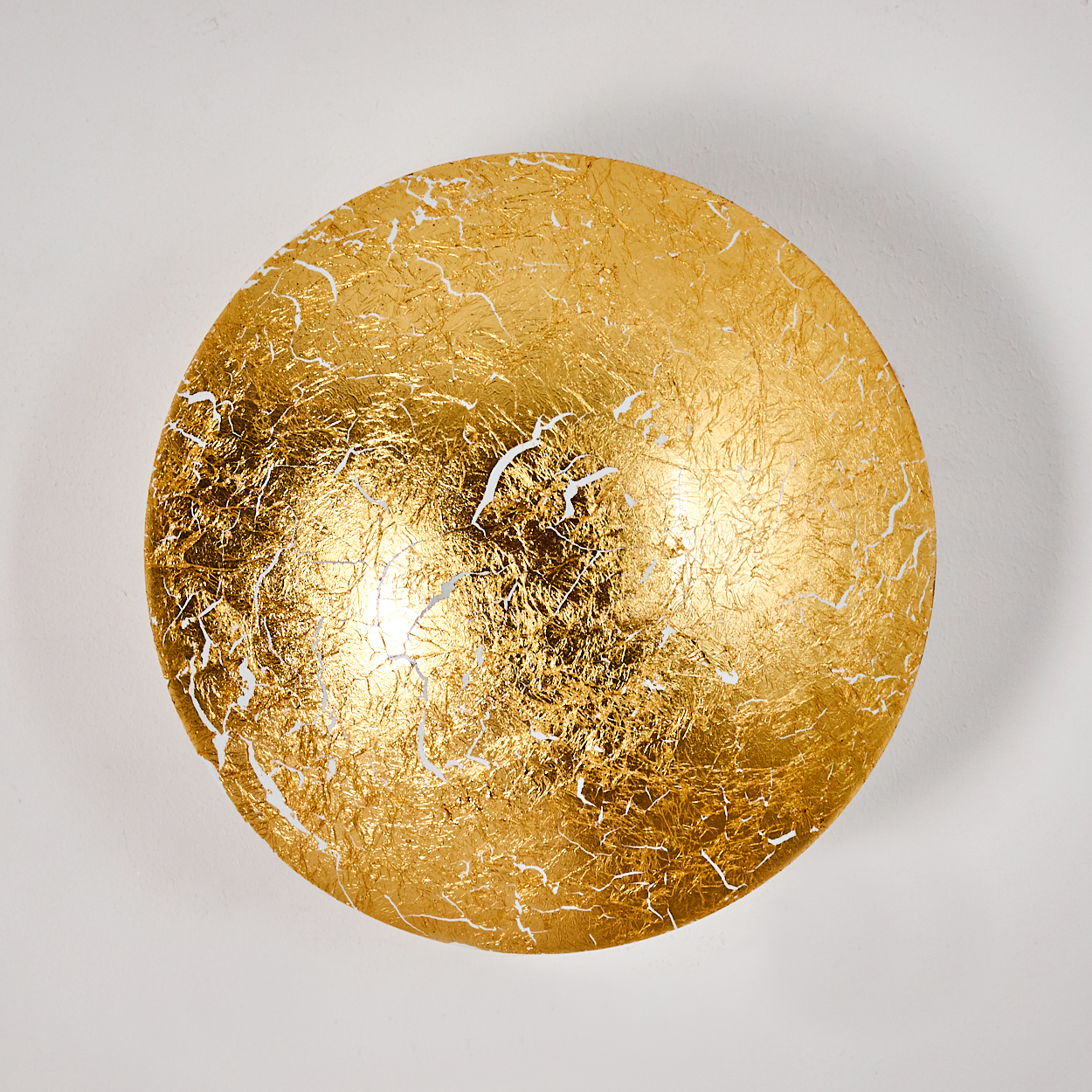 Ein rundes, goldbeschlagenes Objekt ist vor einer weißen Wand zentriert. Risse und Unvollkommenheiten sind auf der Goldoberfläche sichtbar.