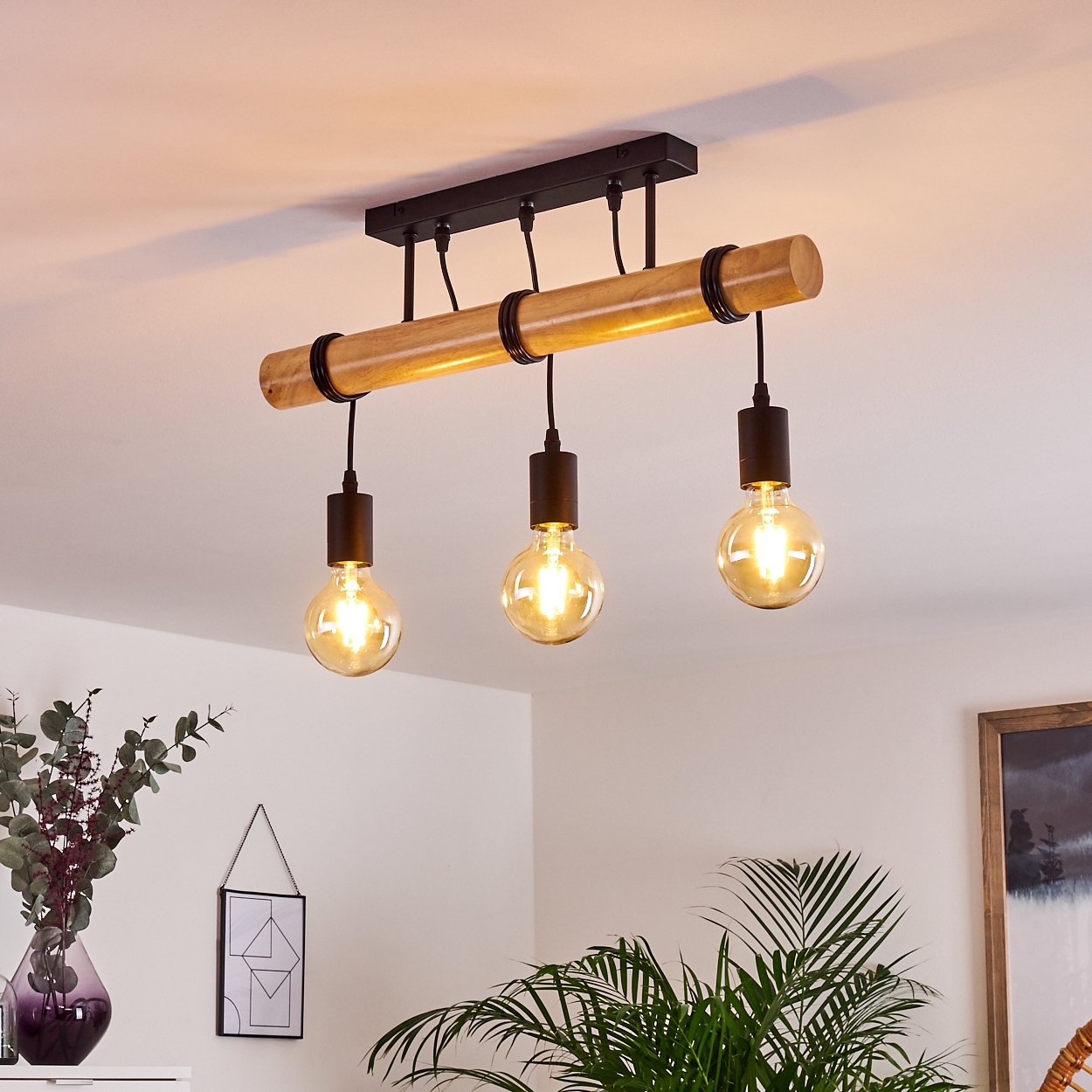 Eine Holz- und schwarze Metallleuchte mit drei Edison-Glühbirnen hängt von der Decke. Der Hintergrund zeigt eine weiße Wand und Pflanzen.