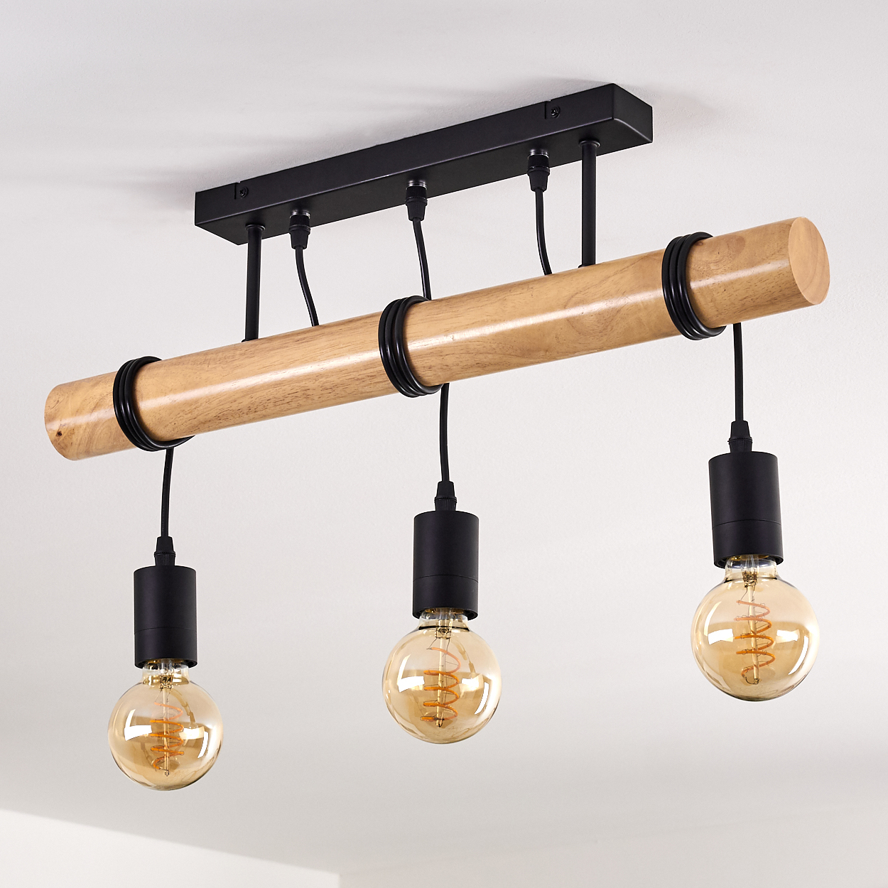 Eine Deckenleuchte mit drei Edison-Glühbirnen. Ein Holzbalken trägt schwarze Halterungen und Glühbirnen. Der Hintergrund ist schlicht weiß.