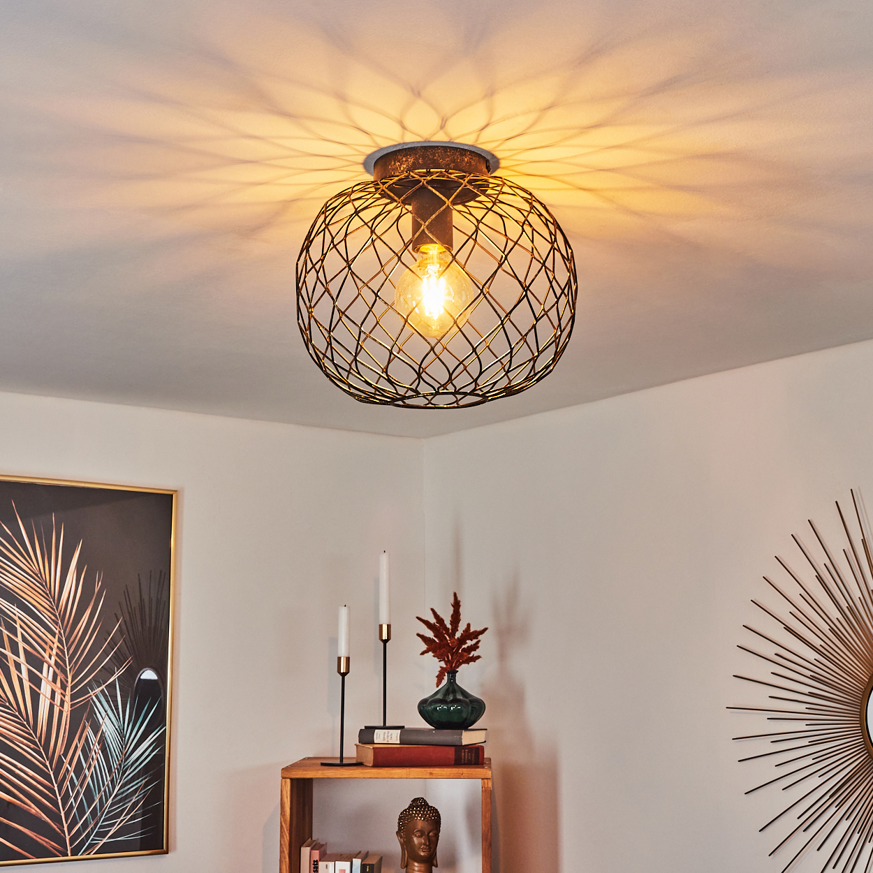 Deckenleuchte mit Drahtkäfig. Darunter ein Regal mit Kerzen, einer Vase und Büchern. Ein gerahmtes Kunstwerk und ein Sonnenstrahlenspiegel an der Wand.