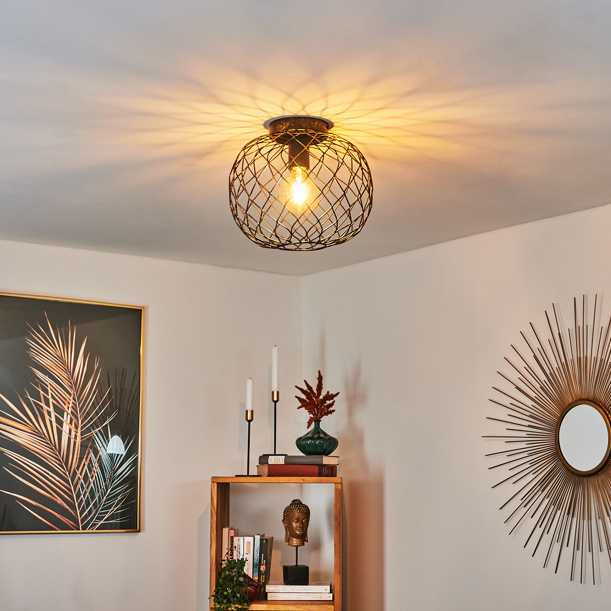 Eine dekorative Leuchte an der Decke. Darunter ein Holzregal mit Kerzen, einer Vase, Büchern und einem Buddha-Kopf. An der Wand ein goldener Spiegel.