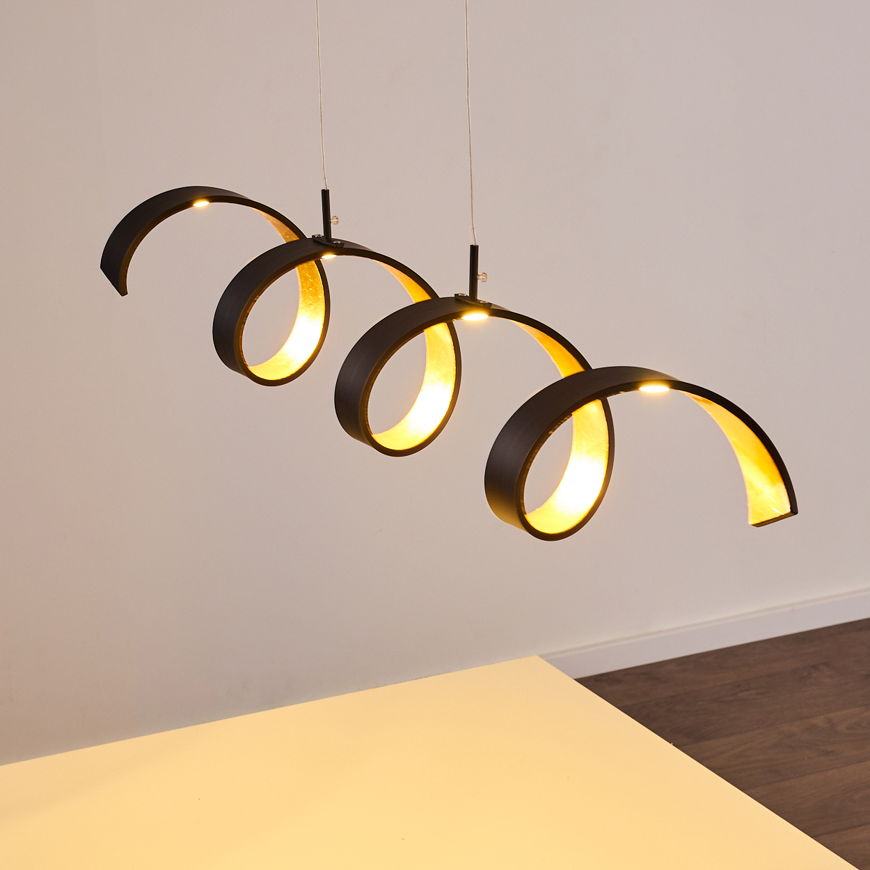 Drei schwarze, spiralförmige Pendelleuchten mit goldenem Innenraum hängen von der Decke. Im Vordergrund befindet sich ein weißer Tisch.