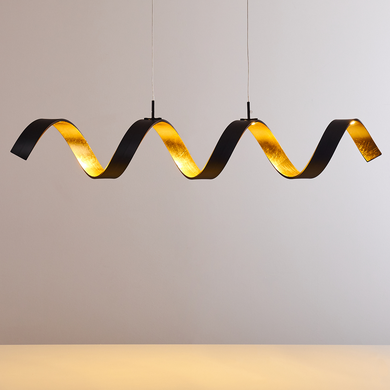 Eine schwarze, wellenförmige Leuchte mit goldener Innenseite hängt vor einem neutralen Hintergrund. Das Licht wird von dünnen Drähten getragen.