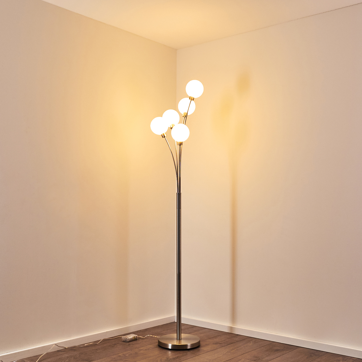Eine moderne Stehlampe mit fünf leuchtenden Kugeln. Sie steht in einer Zimmerecke und wirft Schatten auf beige Wände und Holzboden.