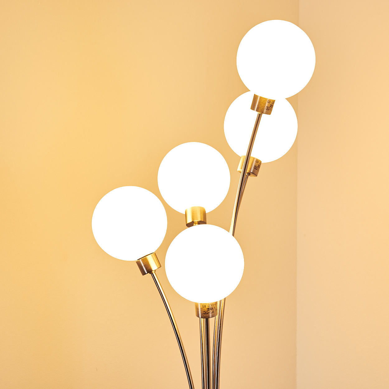 Eine moderne Stehlampe mit fünf weißen Kugelleuchten auf goldenen Stielen, vor einer weichen gelben Wand.