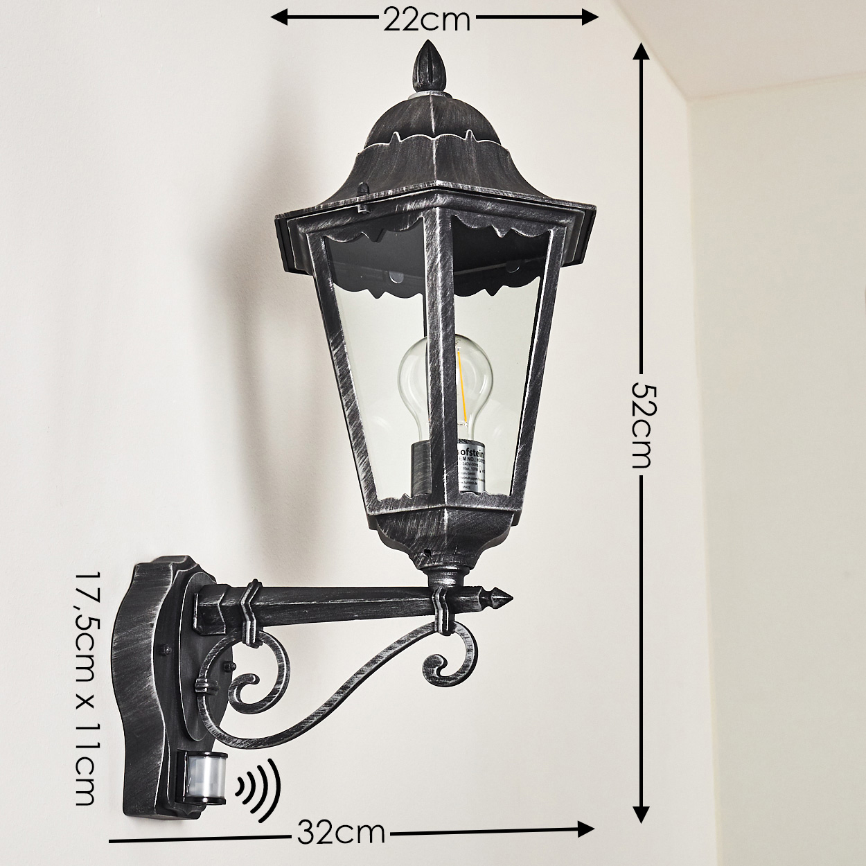 Eine wandmontierte, silberne Außenlampe. Sie hat ein Glasgehäuse für die Glühbirne und einen Bewegungssensor. Abmessungen sind enthalten.
