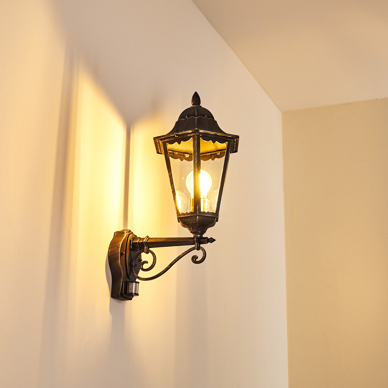 Eine verzierte Wandleuchte mit einer brennenden Glühbirne. Die Lampe ist schwarz und goldfarben und wirft einen warmen Schein auf die beige Wand.