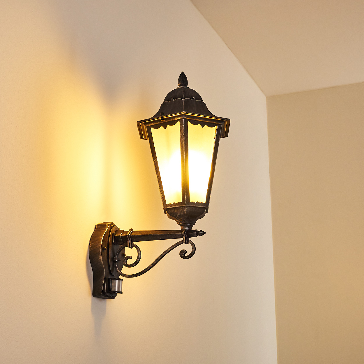 Eine verzierte, schwarze Wandlaterne mit hellem gelbem Licht an einer beigen Wand. Das Licht wirft Schatten.