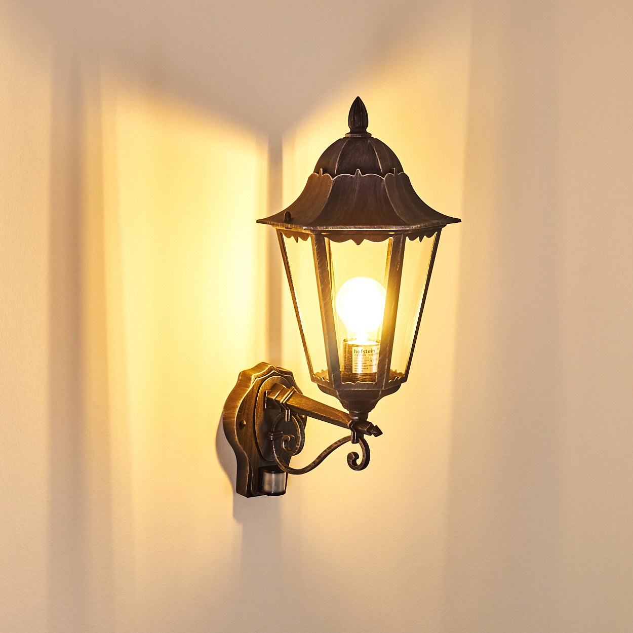 Eine bronzefarbene Wandlampe mit einer leuchtenden Glühbirne ist an einer beigefarbenen Wand montiert. Die Lampe wirft einen warmen Schein.