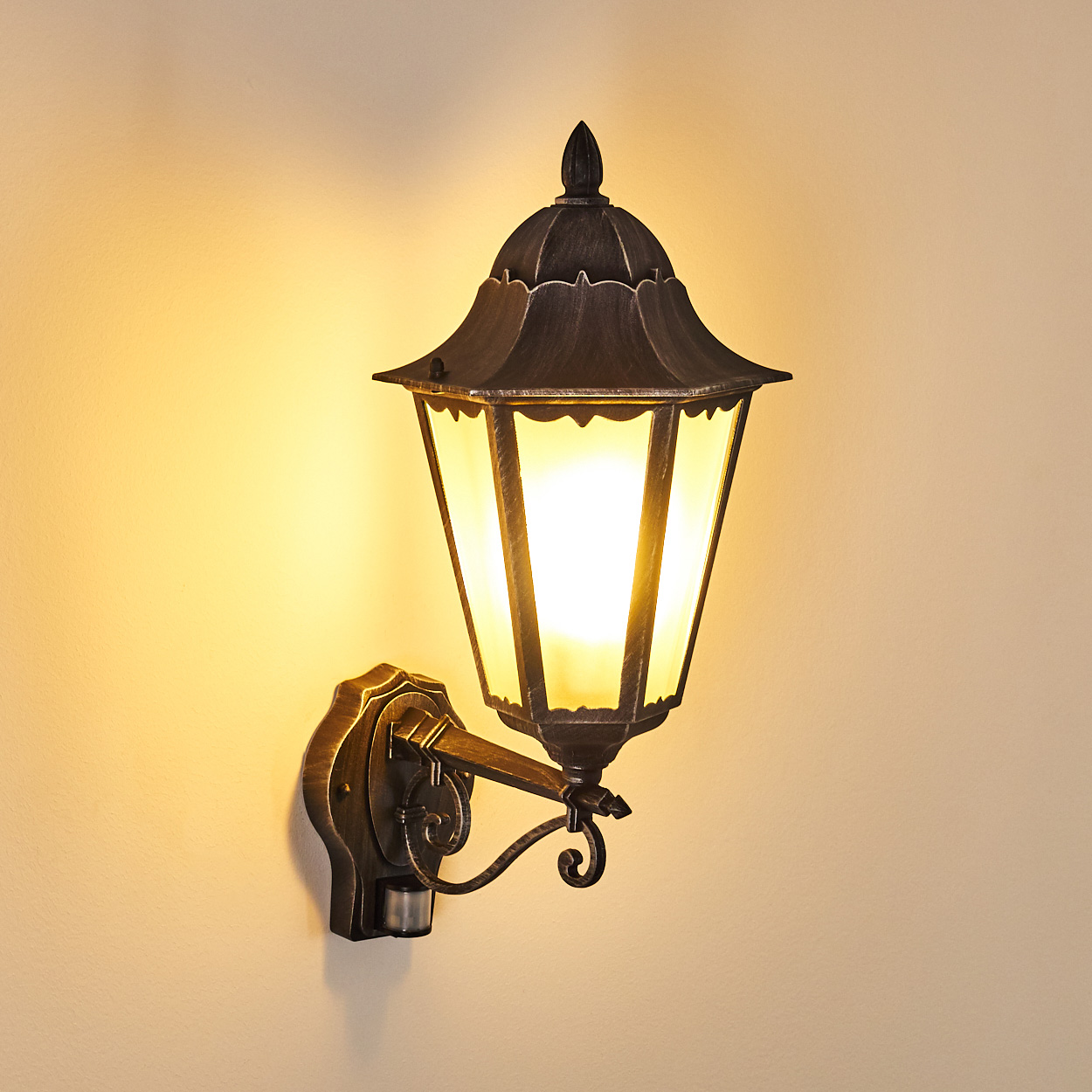 Eine an der Wand befestigte, verzierte Außenlampe mit hellem Licht. Die Lampe ist aus dunklem Metall mit Glasscheiben, an einer hellbeige Wand befestigt.