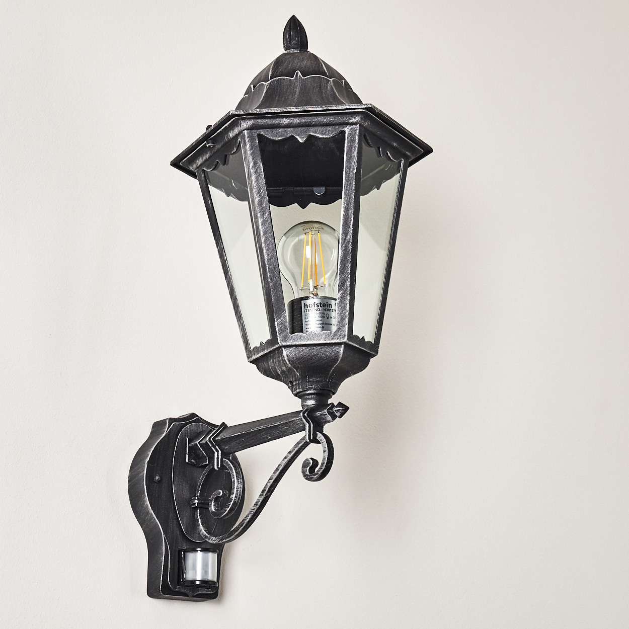 Eine Wand-Außenlampe mit dekorativer Halterung. Die Lampe hat ein Glasgehäuse und eine Edison-Glühbirne. Sie ist an einer hellen Wand befestigt.