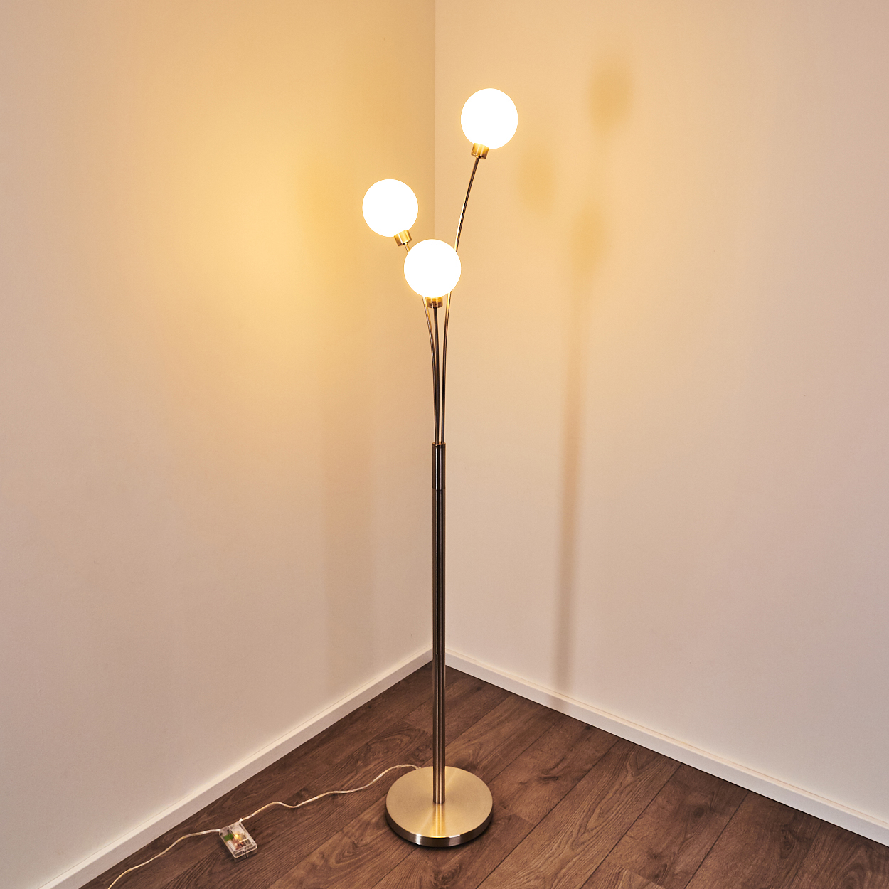 Eine goldene Stehlampe mit drei kugelförmigen Lichtern steht in einer Zimmerecke. Der Raum hat Holzböden und weiße Wände.