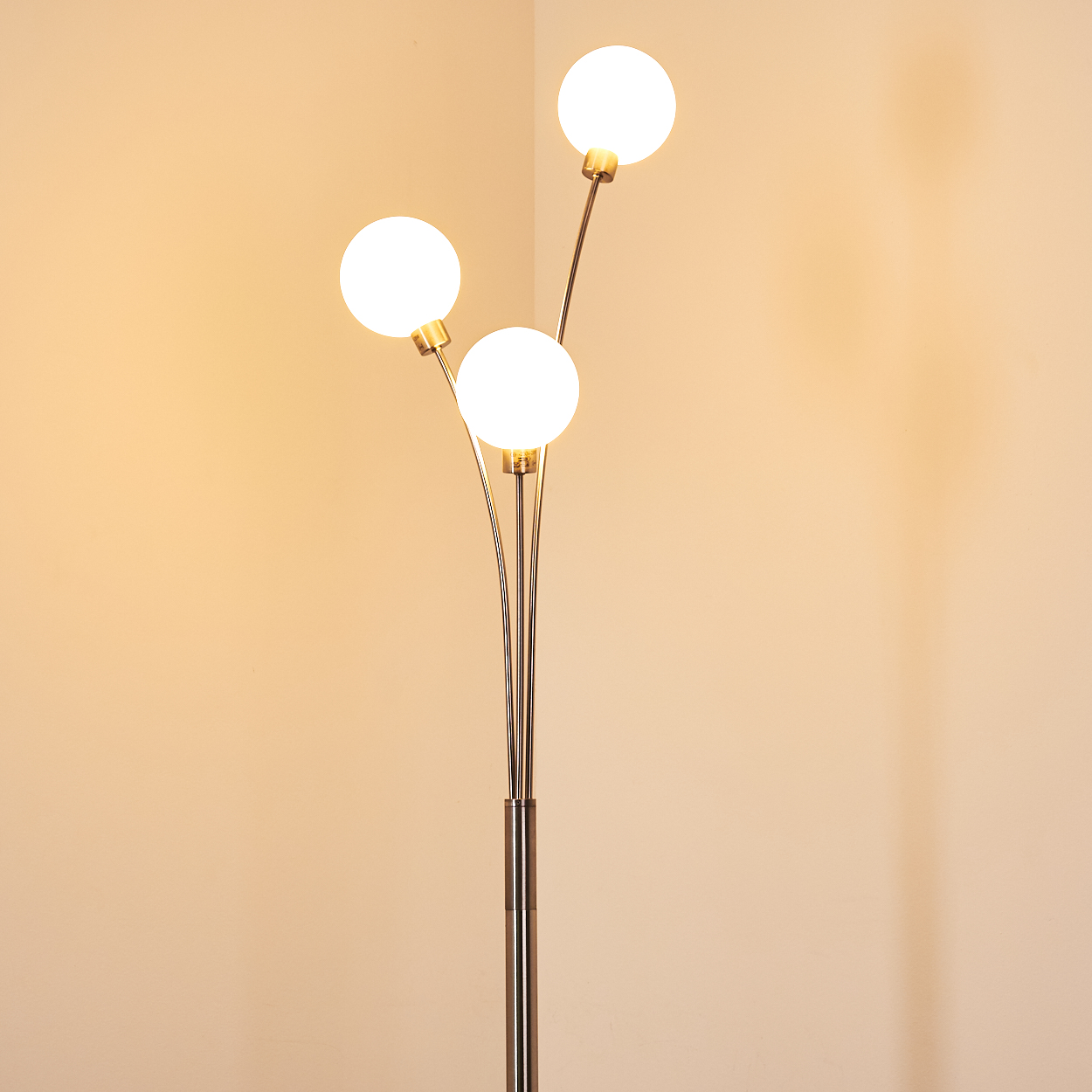 Eine Stehlampe mit drei kugelförmigen weißen Lichtern. Die Lampe hat einen silbernen Sockel und goldene Stiele, vor einer cremefarbenen Wand.