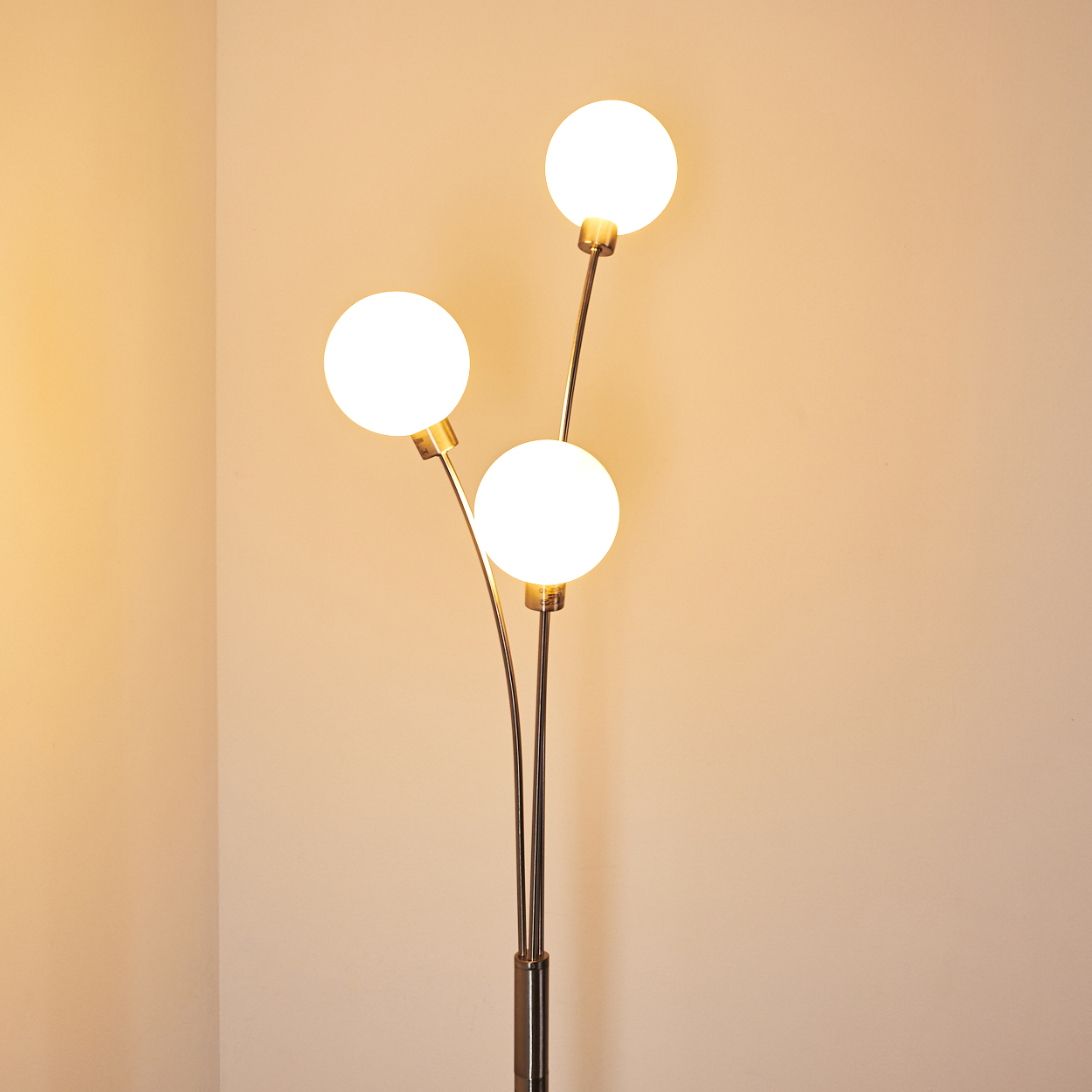 Eine Stehlampe mit drei weißen kugelförmigen Glühbirnen. Die Lampe hat einen metallischen Stiel und steht vor einer hellbeigen Wand.