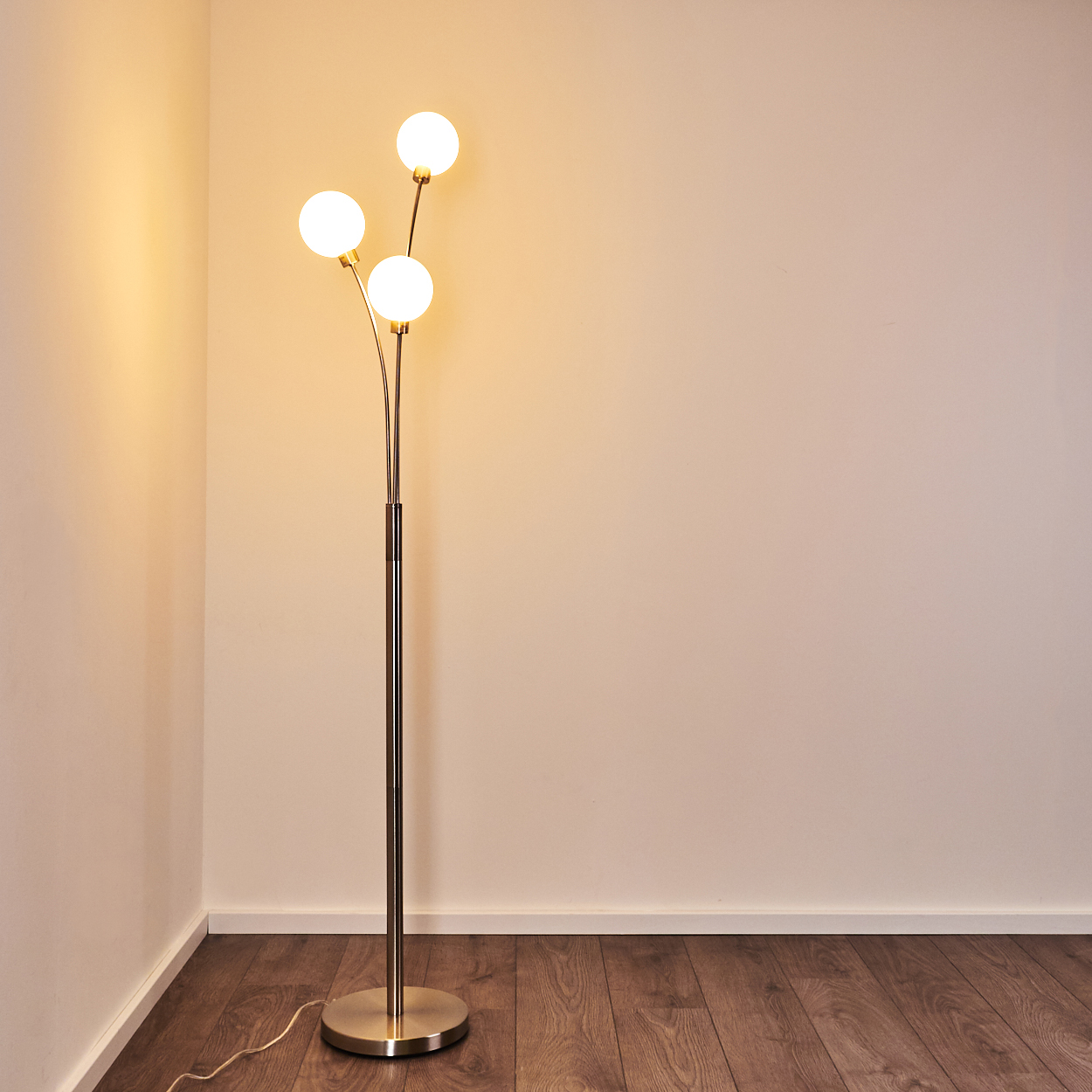 Eine Stehlampe mit drei kugelförmigen Lichtern steht in einer Ecke. Die Lampe ist goldfarben. Der Hintergrund ist beige Wände und Holzboden.