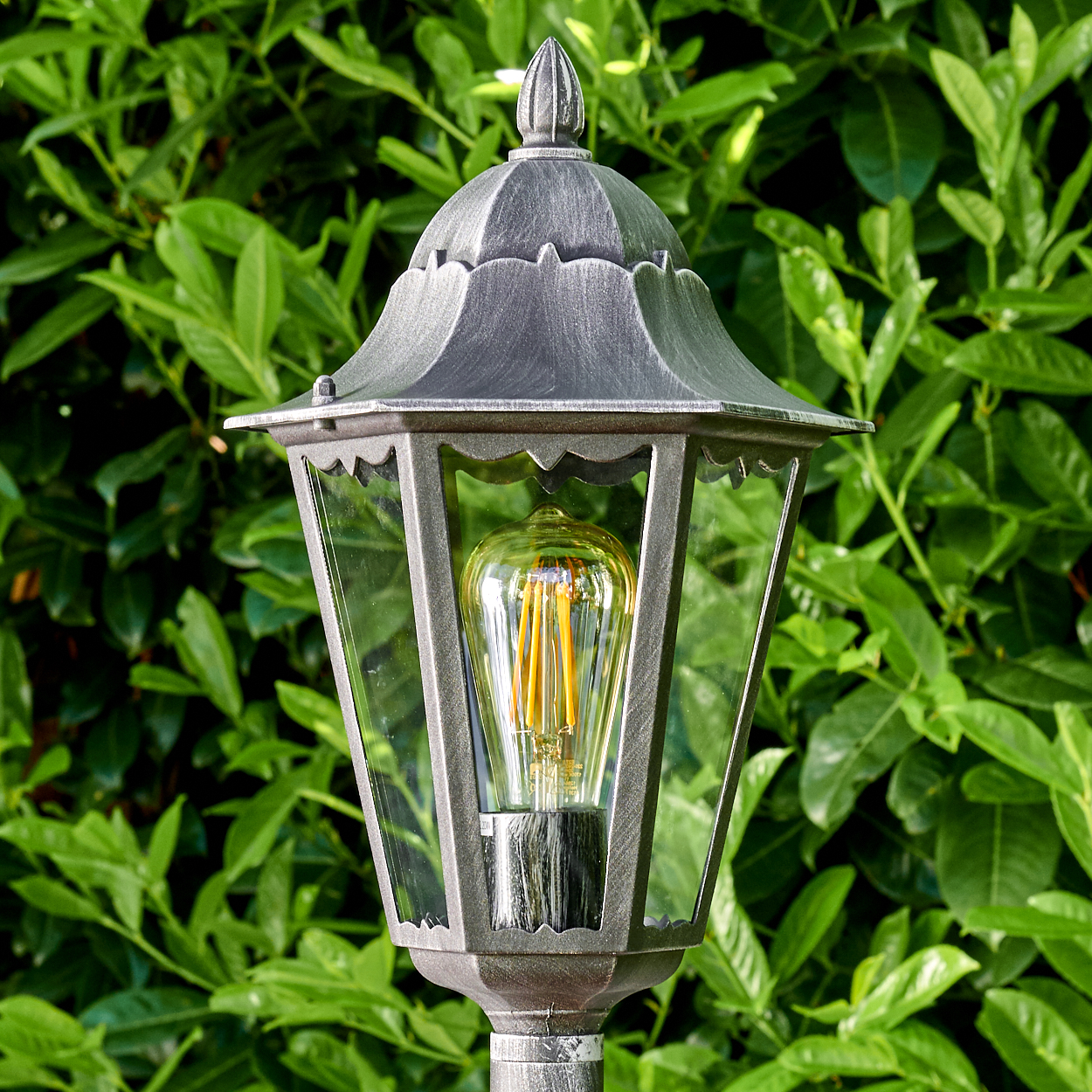 Eine graue Außenlampe mit dekorativer Glühbirne steht vor grünem Laub. Die Lampe hat ein klassisches Design.