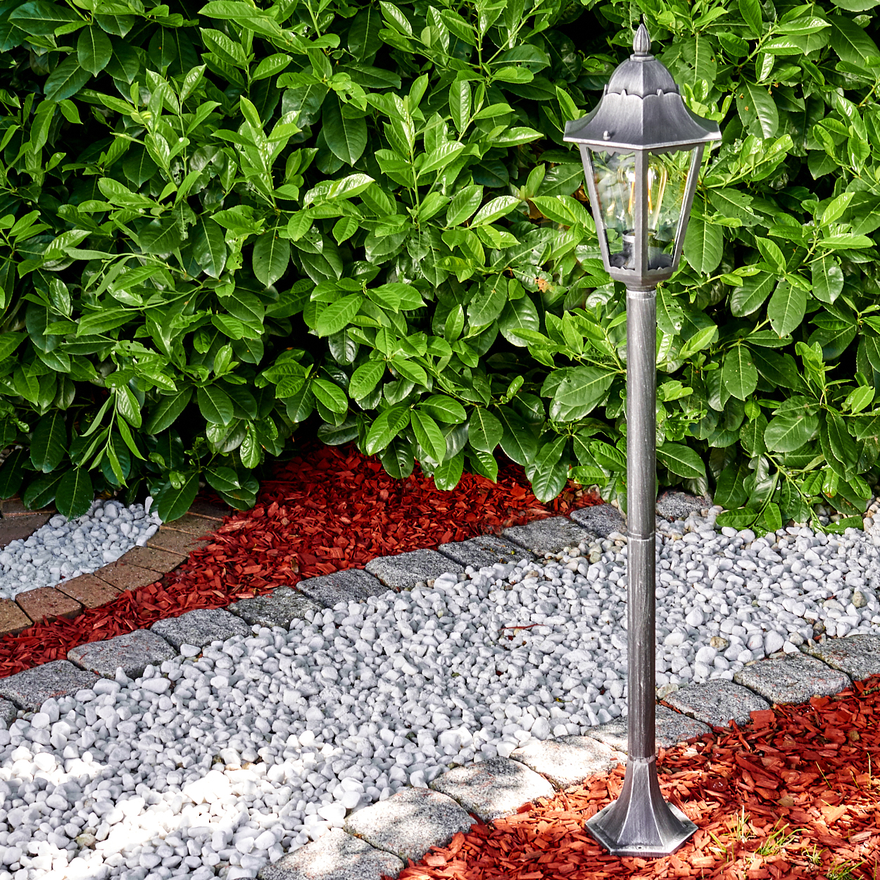 Eine Gartenszene mit Laternenpfahl, weißen Steinen, roter Mulche und grünen Büschen im Hintergrund.