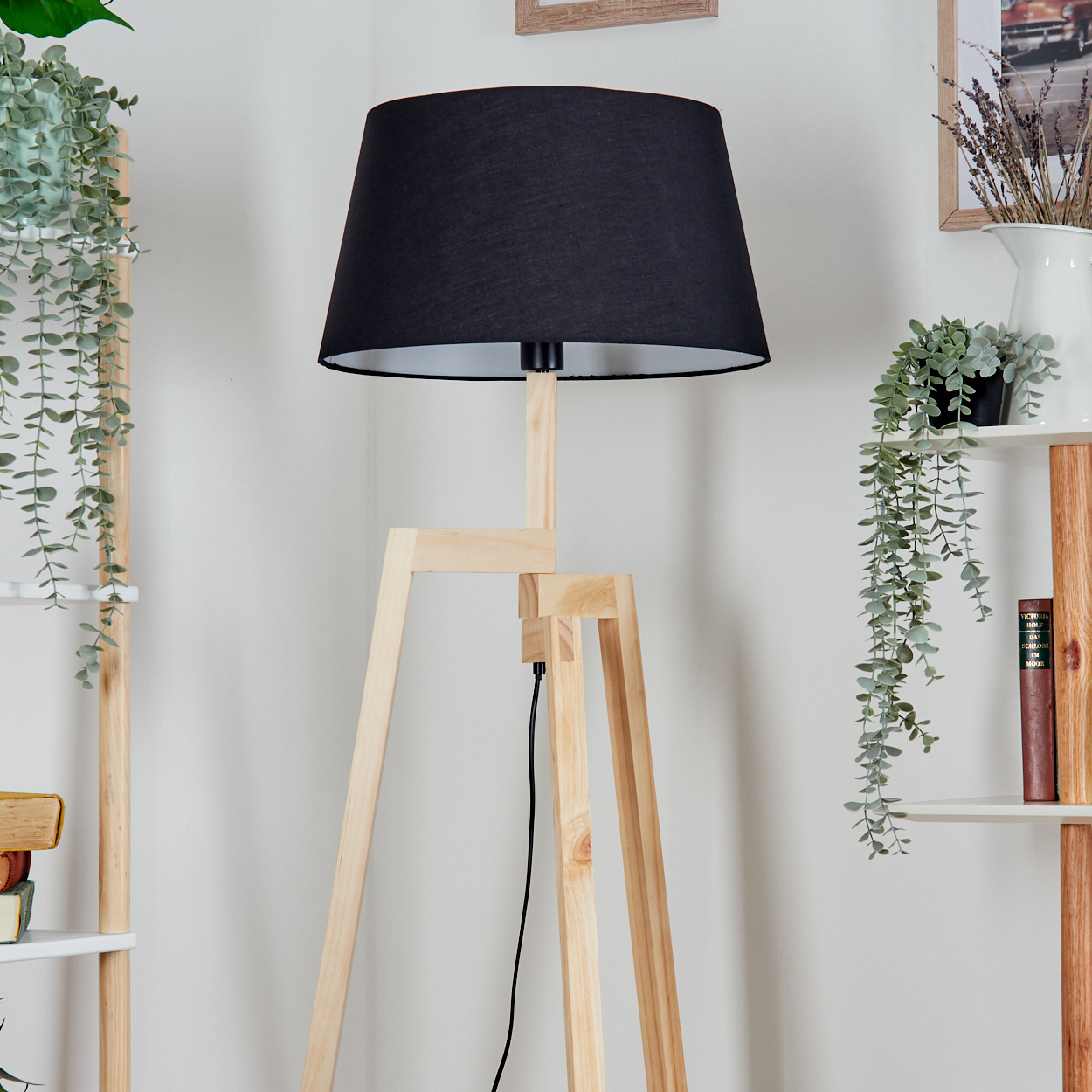 Eine Stehlampe aus Holz mit schwarzem Lampenschirm steht in einem Raum. Regale mit Pflanzen und Büchern sind im Hintergrund zu sehen.