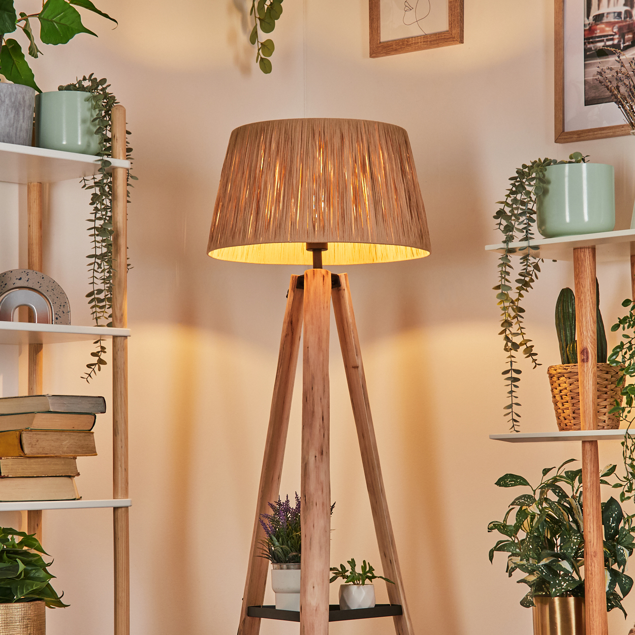 Eine Holz-Dreibein-Lampe mit gewebtem Schirm steht im Mittelpunkt. Regale mit Pflanzen und Büchern sind beidseitig. Warmes Licht.