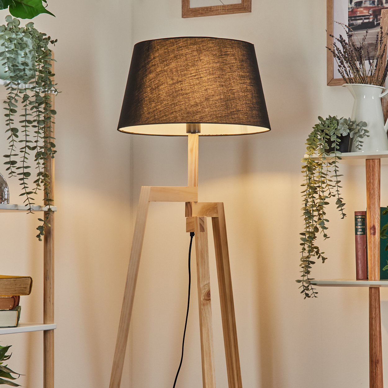 Eine hölzerne Stehlampe mit schwarzem Lampenschirm. Regale mit Pflanzen und Büchern befinden sich auf beiden Seiten der Lampe.
