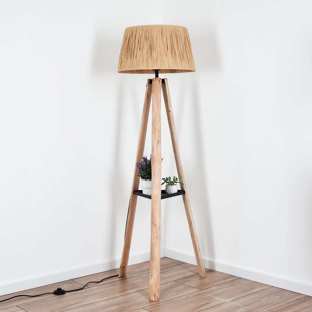 Eine hölzerne Stehlampe mit Web-Schirm. Sie hat ein kleines Regal mit zwei Topfpflanzen. Die Lampe steht in einer Ecke.