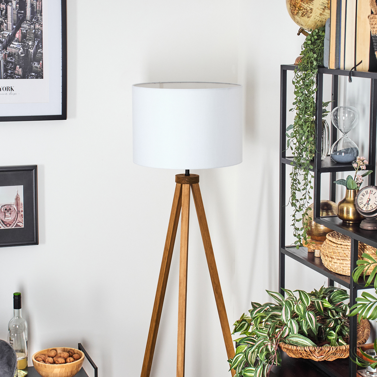 Eine hölzerne Stativlampe mit weißem Schirm steht neben einem schwarzen Regal mit Dekoration. An der Wand hängt ein gerahmtes New York-Bild.