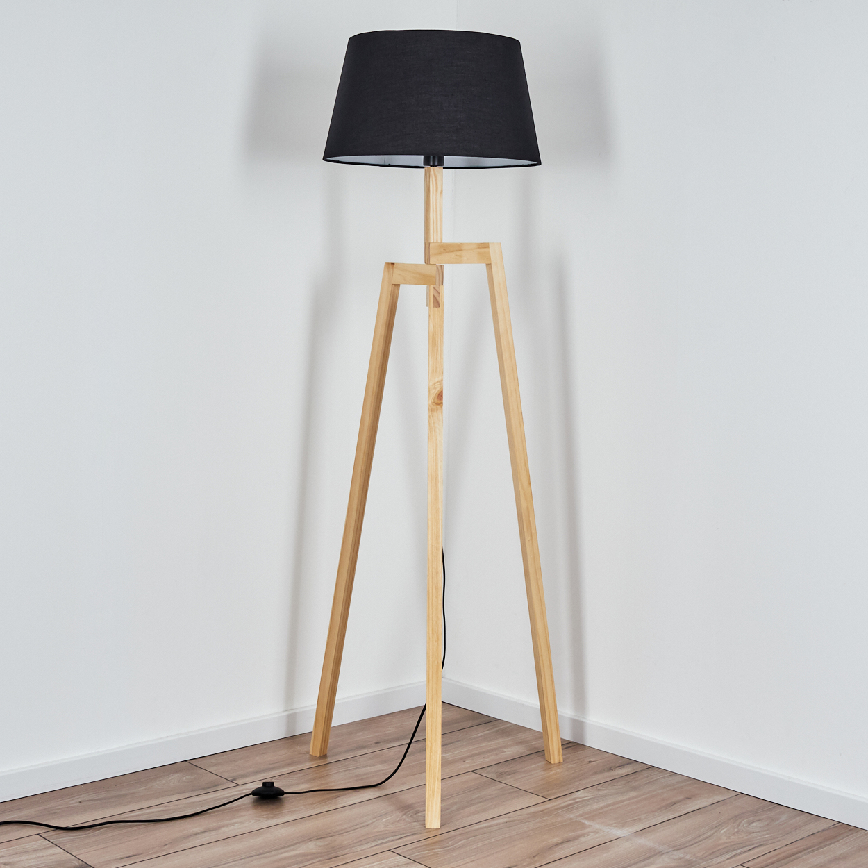 Eine Holz-Stativ-Stehlampe mit schwarzem Lampenschirm steht in einer Zimmerecke. Der Boden ist aus Holz, die Wände sind weiß.