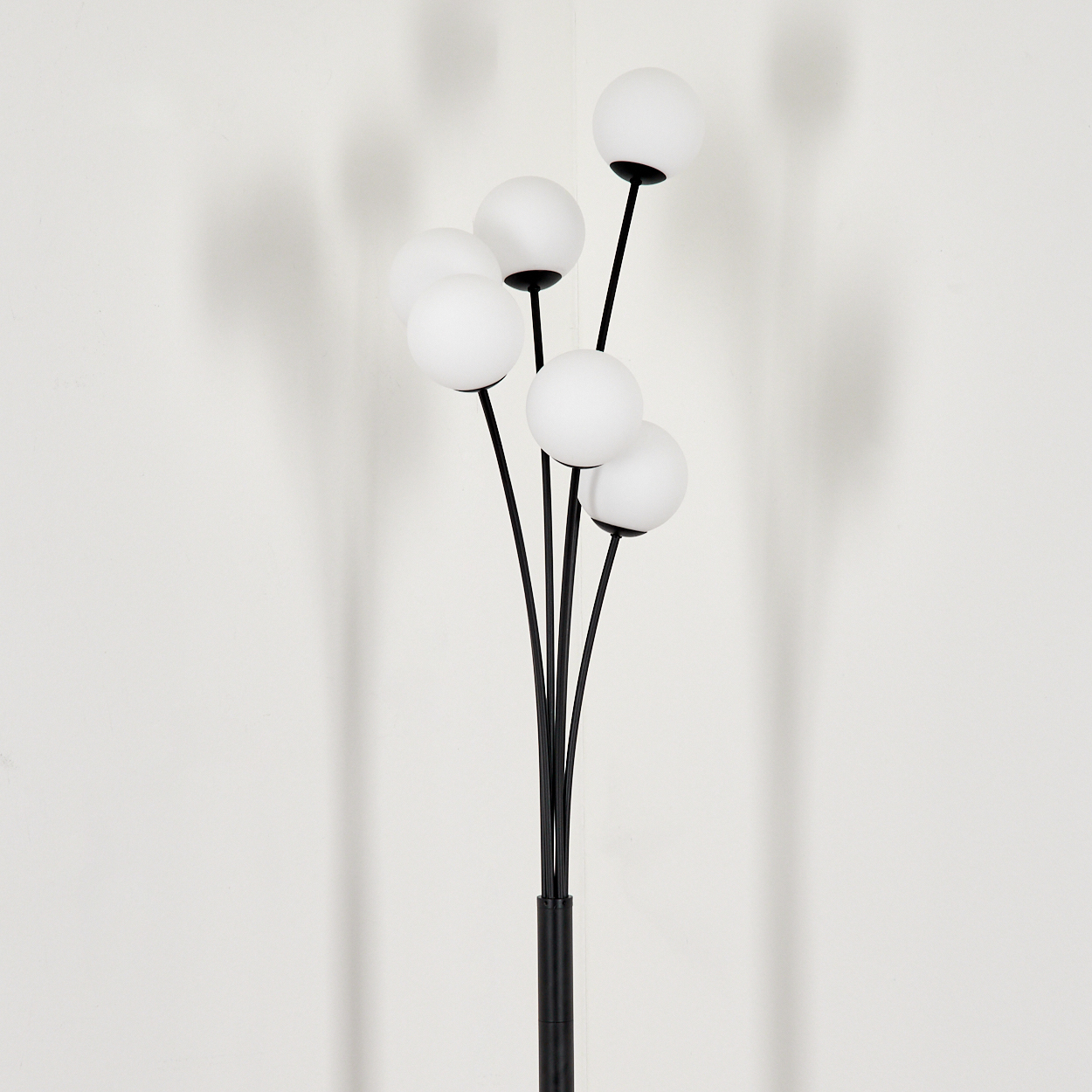 Eine moderne Stehlampe mit schwarzem Fuß und Stielen, gekrönt von weißen Kugelleuchten vor einer weißen Wand mit Schatten.