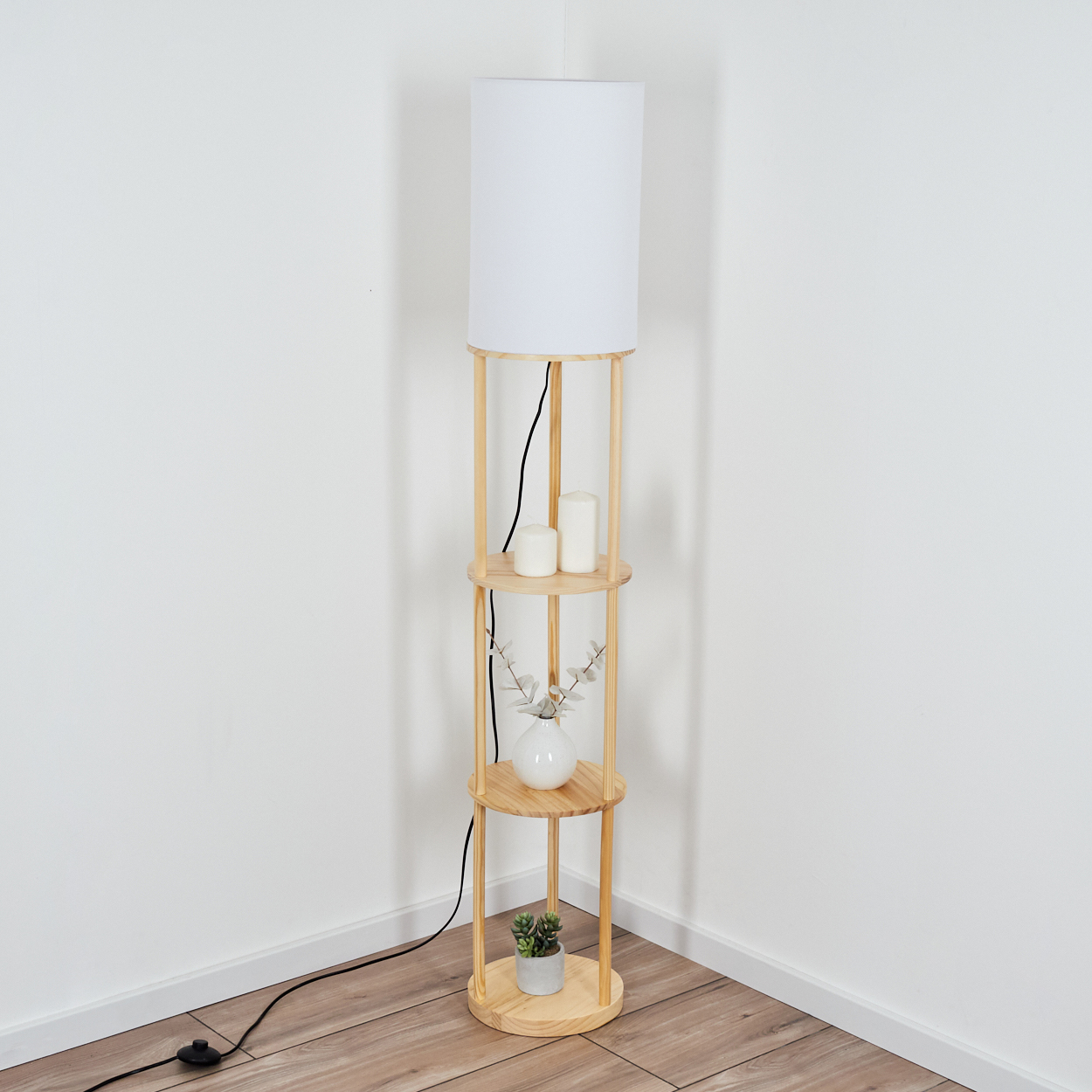 Eine Stehlampe aus Holz mit weißem Lampenschirm und Regalen mit Kerzen, einer Vase und einer Pflanze vor weißer Wand.