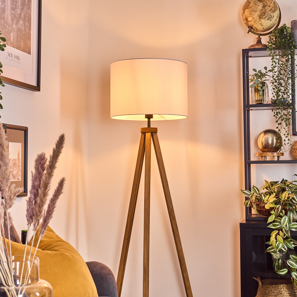 Eine beleuchtete Lampe mit weißem Schirm steht auf Holzbeinen. Im Hintergrund ein Regal mit Pflanzen und einem Globus. Links eine Couch.