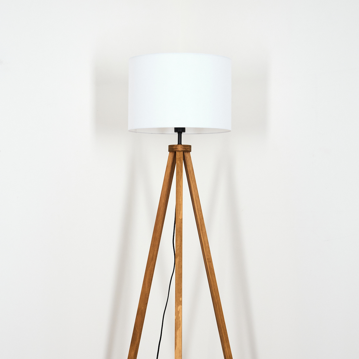 Eine Stehlampe aus Holz mit weißem Lampenschirm steht vor einer weißen Wand. Ein schwarzes Netzkabel verläuft am Bein der Lampe.