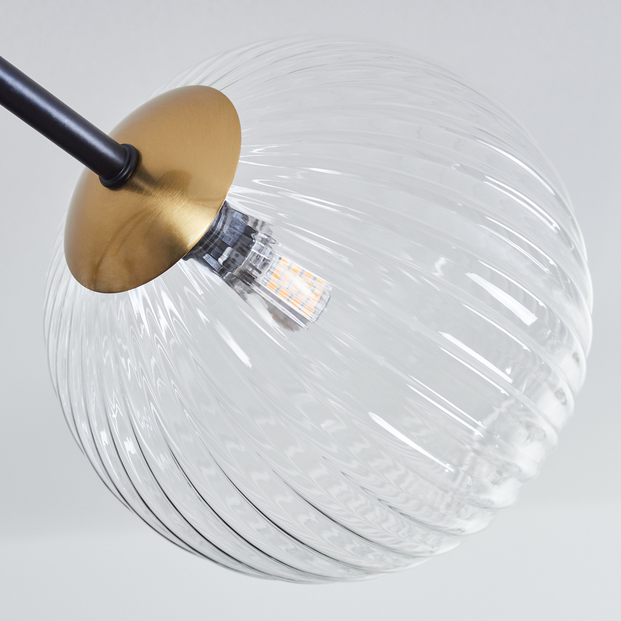 Nahaufnahme einer Glaskugel-Leuchte. Sie hat eine goldene Kappe, einen schwarzen Arm und eine klare, gewirbelte Glaskugel mit LED-Lampe.