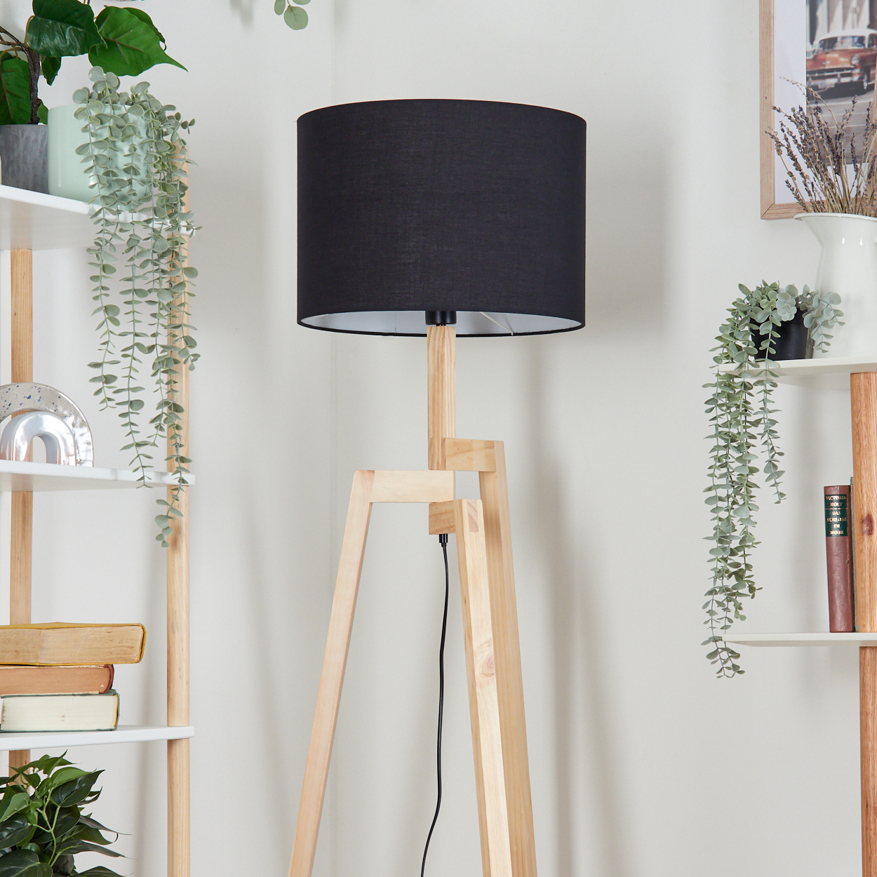 Eine Stehlampe aus Holz mit schwarzem Lampenschirm steht vor einer weißen Wand. Regale mit Pflanzen befinden sich an den Seiten.