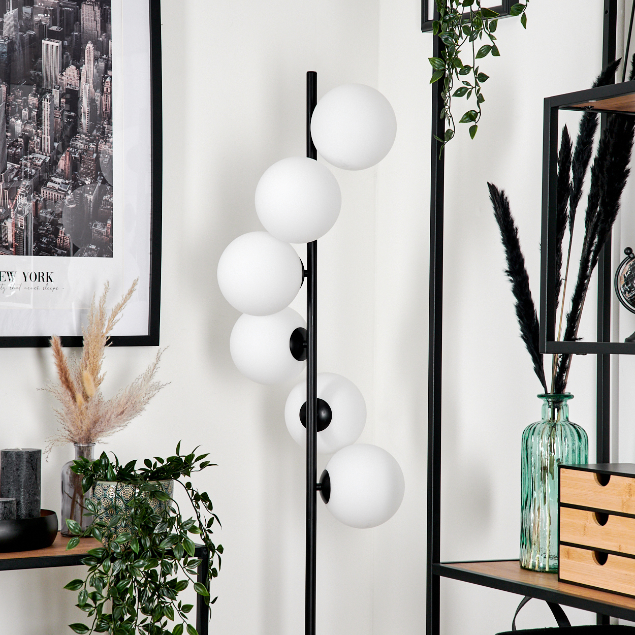 Ein modernes Interieur mit einer schwarz-weißen Lampe mit kugelförmigen Lichtern. Regale halten Pflanzen und dekorative Gegenstände. Ein New York-Druck hängt.